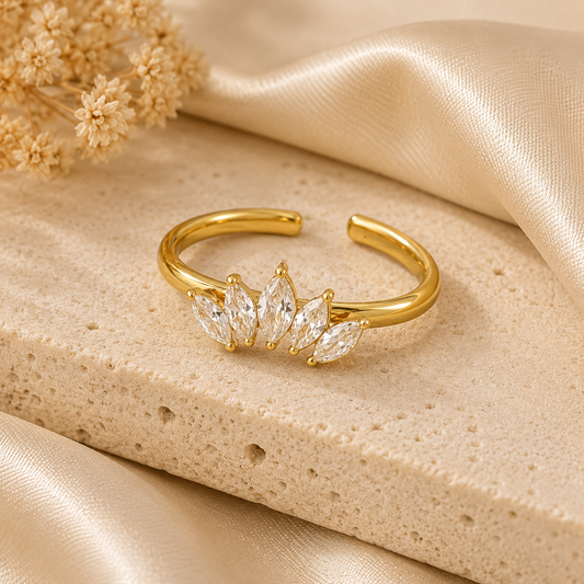 Gold Radiance Ring featuring five marquise-cut crystal stones in a crown-inspired design, displayed on a neutral stone surface with soft satin fabric and warm natural lighting.